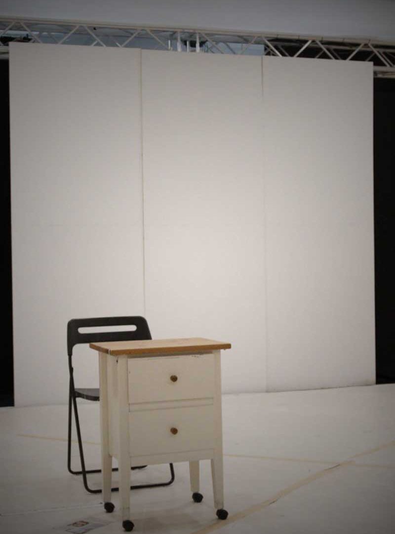 An empty bright rehearsal room with a black chair and a small white chest of drawers