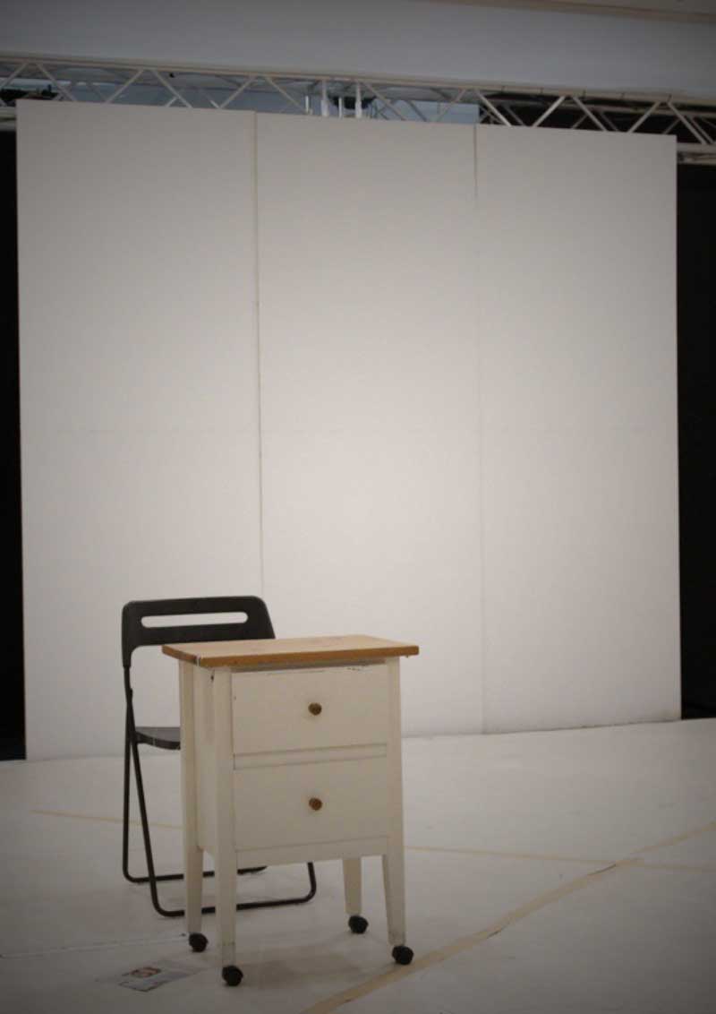 An empty bright rehearsal room with a black chair and a small white chest of drawers