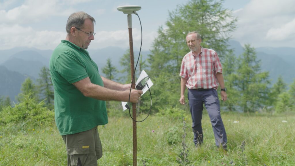 Sterben-20-redmonster_Pax-Almbestattung-GPS Vermessung-2 Two men on an alpine meadow surveying a plot with a GPS pole, larch trees and mountains behind them.