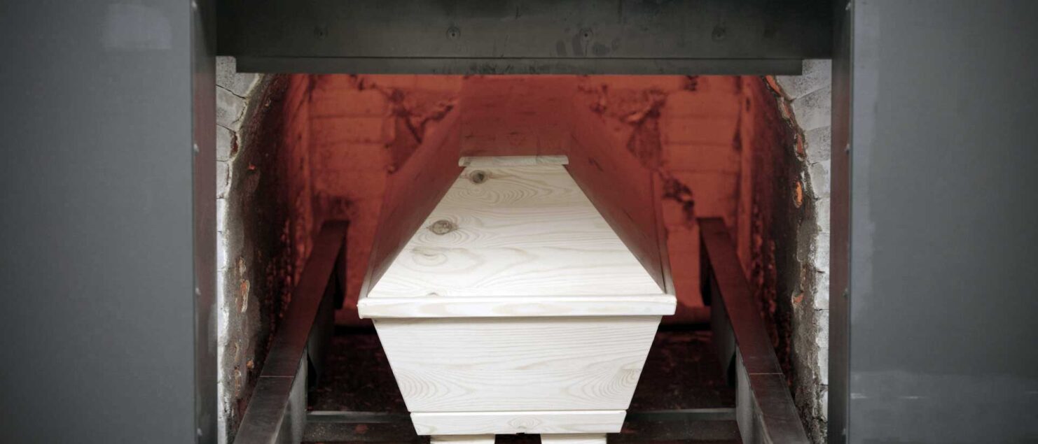 Ein heller Holzsarg steht in der geöffneten Brennkammer eines Krematoriums, die Schamottwände glühen rot.