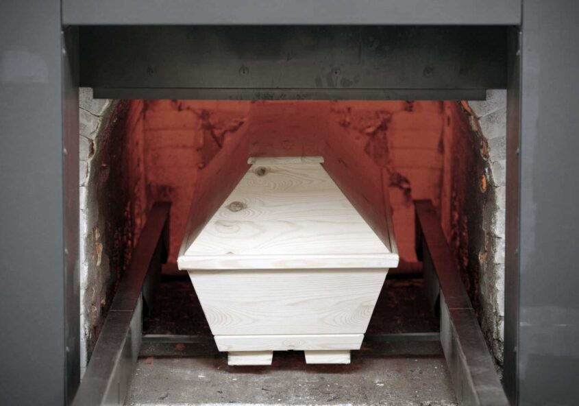 Ein heller Holzsarg steht in der geöffneten Brennkammer eines Krematoriums, die Schamottwände glühen rot.