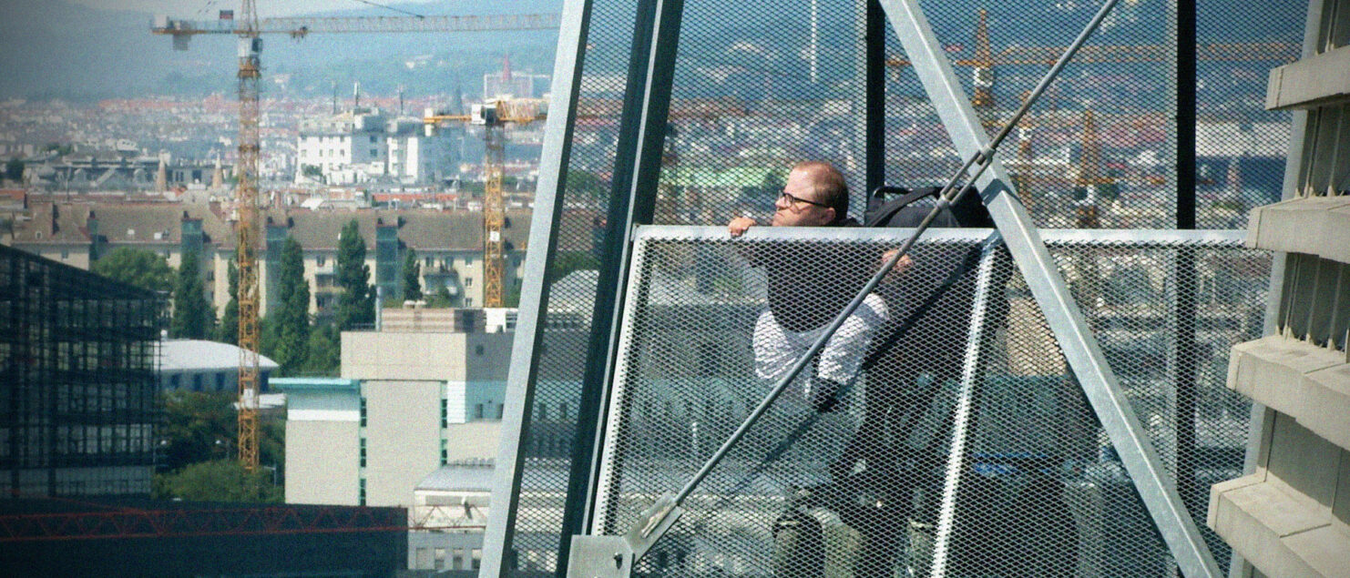 Ein Mann im Elektrorollstuhl auf einem Steg hoch über Wien, im Hintergrund Baukräne.