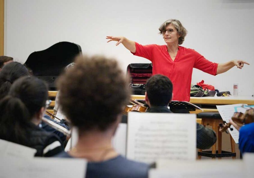 Eine Dirigentin in Rot vor einem Orchester, im Vordergrund Musizierende mit Noten.