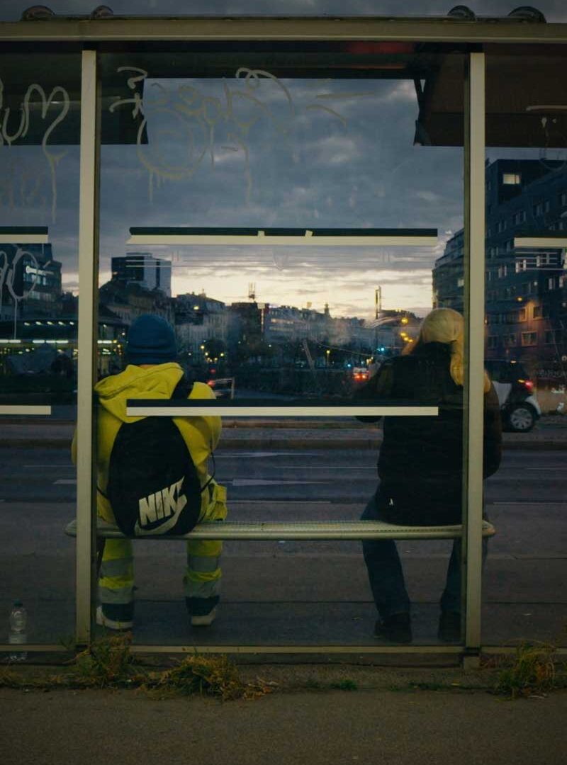 Zwei Menschen sitzen von hinten gesehen in einem Wartehäuschen, einer in gelber Warnjacke, dahinter die Stadt in der Dämmerung.