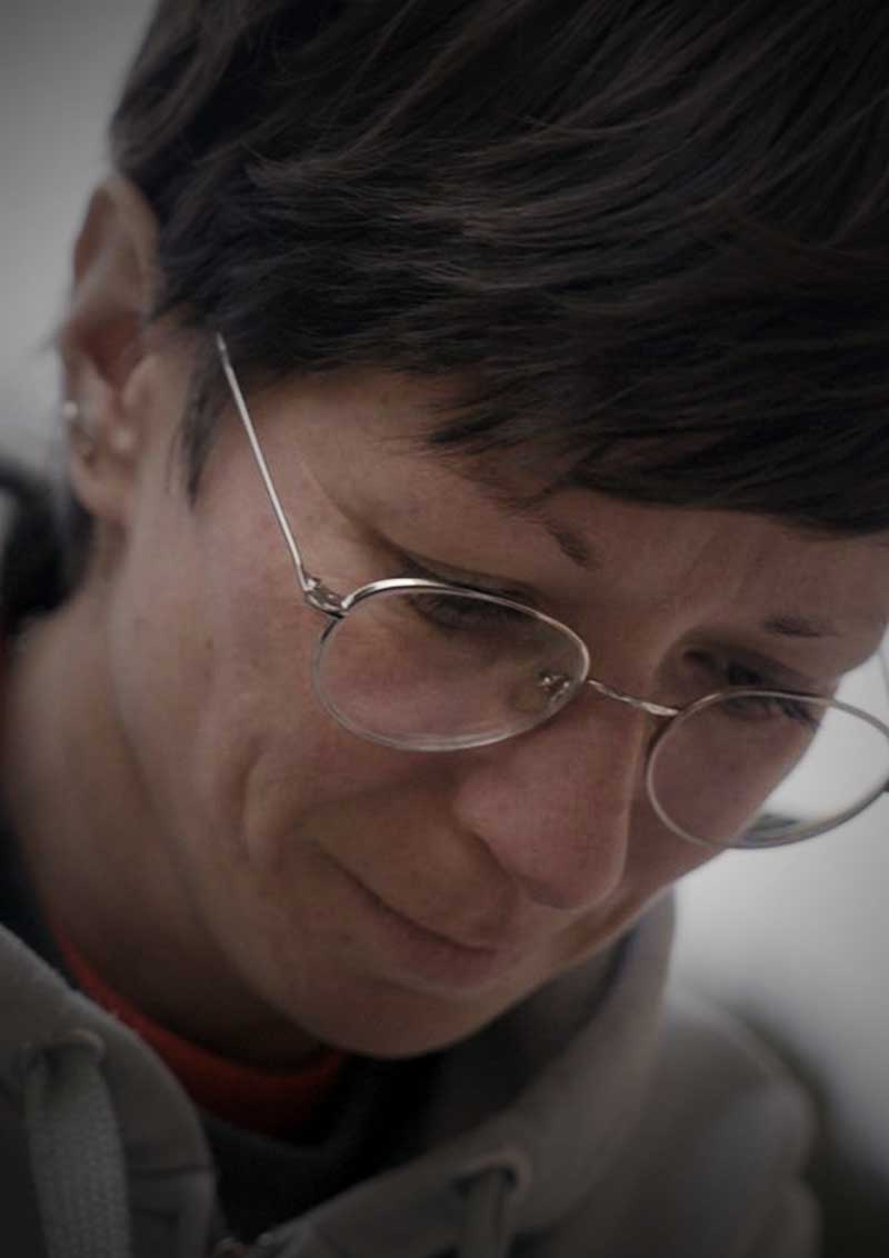 Nahaufnahme einer Frau mit kurzen dunklen Haaren und feiner Metallbrille, die den Blick senkt und leicht lächelt.