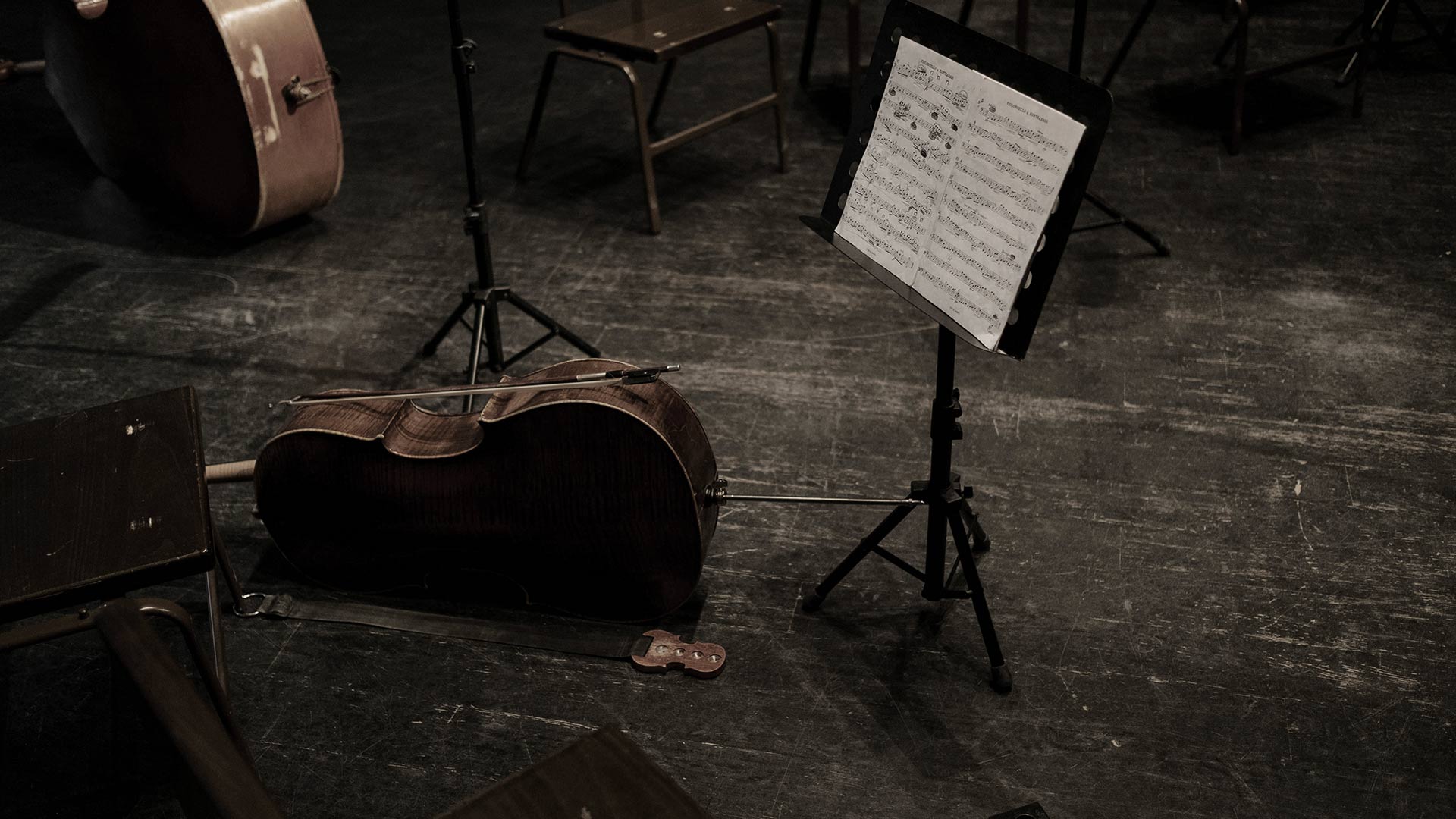 Geigenkasten und Notenständer in einem leeren Proberaum. Holzstühle, dunkler Boden.