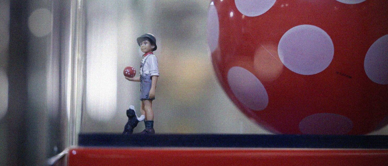 Eine kleine Spielzeugfigur mit rotem Ball steht neben einem großen rot weiß gepunkteten Ball in einer Vitrine.