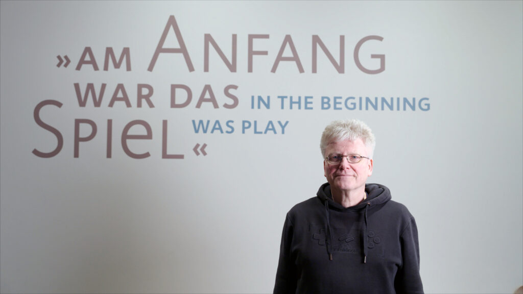 Homo Ludens – Still 01 Jens Junge, founder of the Institute for Ludology, in front of the lettering “In the beginning was play”