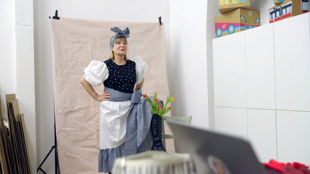 Nicht jung, nicht leise – Still 03 Frau im Kostüm mit Schürze und Kopftuchschleife vor einem Fotohintergrund