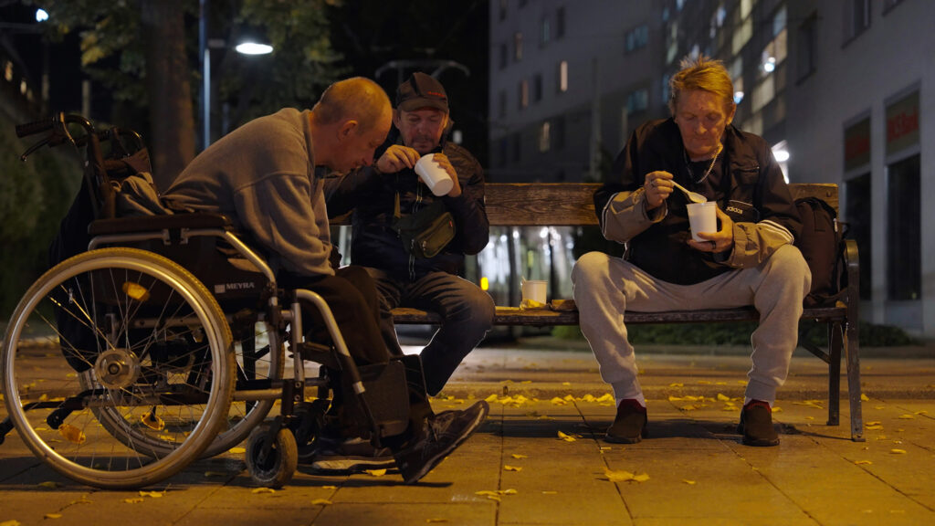 Three men eating soup from the Canisibus on a park bench at night