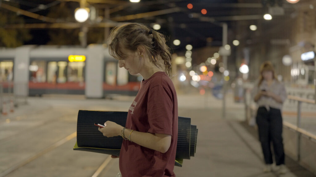 Veronika Müller at a tram stop at night, looking at her phone