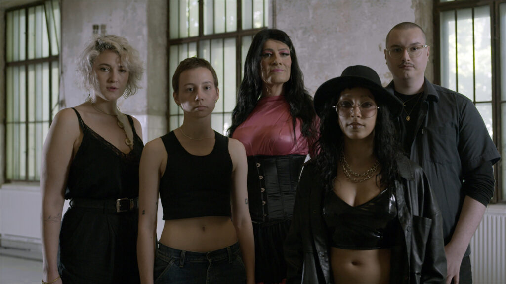 Beyond Binaries – still 1 Five people stand side by side in a high-ceilinged room with tall windows, looking into the camera.