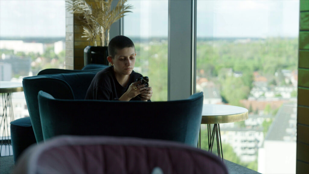Beyond Binaries – still 2 A person with closely cropped hair sits in an armchair by a floor-to-ceiling window high above the city, looking at their phone.