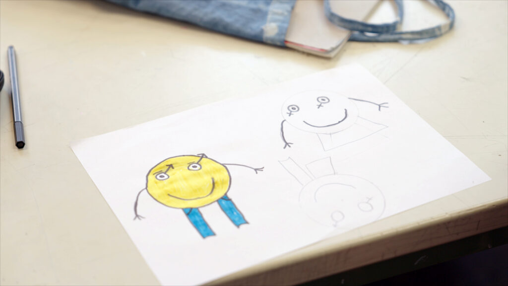 Beyond Binaries – still 3 A child's drawing on a table next to a pen and a pencil case, showing two figures with gender symbols.